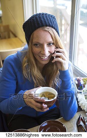 Stock Photograph - Young woman enjoying a cup of tea while talking on her smart phone in a cafe, South-central Alaska; Anchorage, Alaska, United States of America. Fotosearch