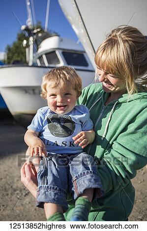 Stock Photograph - Young woman holding her toddler with a boat in the background, South-central Alaska; Homer, Alaska, United States of America. Fotosearch