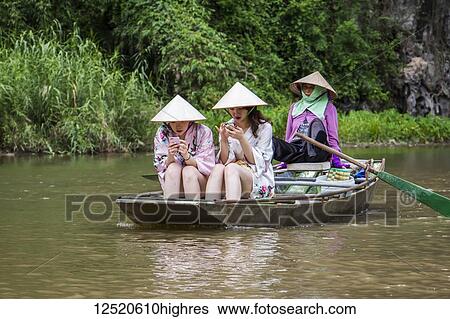 Stock Photograph - Young women in a boat using their cell phones on the Ngo Dong River surrounded by the limestone karst mountains; Tam Coc, Ninh Binh, Vietnam. Fotosearch