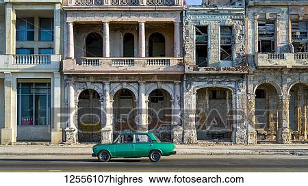 Stock Photograph - An old car passes the weathered facade of old buildings along a street; Havana, Cuba. Fotosearch