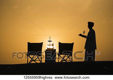 Stock Photograph - Butler holding champagne glasses after setting up a table for a sundowner drink; Rajasthan, India. Fotosearch
