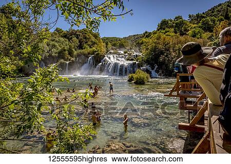 Stock Photograph - People swimming in the Krka River at the Krka waterfalls with tourists standing on the bridge looking out, Krka National Park; Croatia. Fotosearch