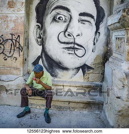 Stock Photograph - Portrait of a man painted on a stone wall along a street in Havana with a senior man sitting on a step eating; Havana, Cuba. Fotosearch