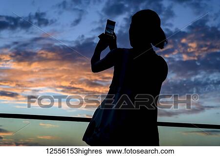 Stock Photograph - Silhouette of a woman photographing the sunset with a smart phone; Havana, Cuba. Fotosearch