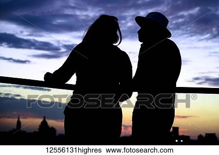 Stock Photograph - Silhouetted couple stands at a railing overlooking the skyline of Havana at sunset; Havana, Cuba. Fotosearch
