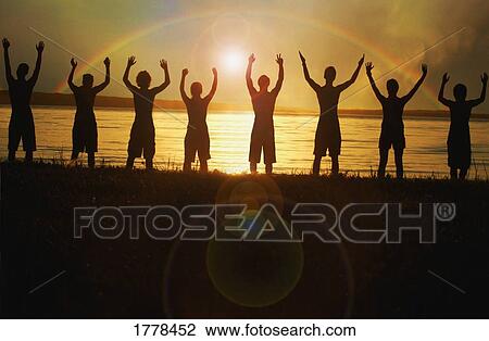 Stock Image - Rainbow behind line of men in water with arms raised. Fotosearch
