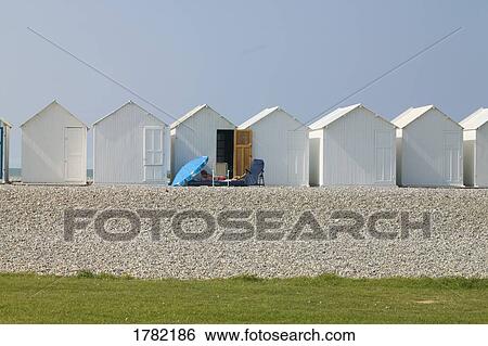 Stock Photograph - A blue umbrella and lounge chair in front of white beach cabins on Cayeux sur-Mer, France. Fotosearch