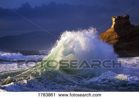 Stock Image - Wave crashing, distant storm, rock cliffs of Cape Kiwanda. Fotosearch
