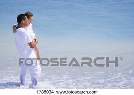 Picture - A father holds his child while standing in the ocean. Fotosearch