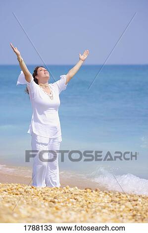 Stock Image - A woman raises her hands at the beach. Fotosearch