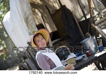 Stock Image - Woman in old-fashioned clothing beside covered wagon. Fotosearch