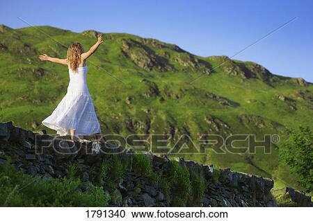 Stock Image - Young woman balancing on rock wall ledge. Fotosearch