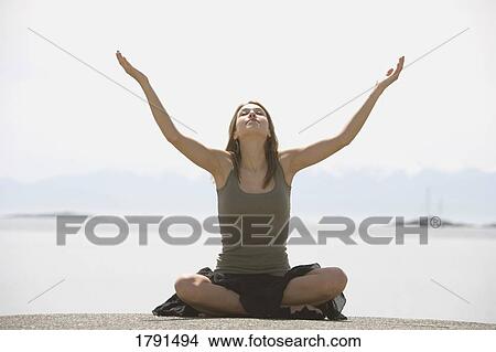Picture - Young woman sitting cross-legged with arms raised in front of beachfront. Fotosearch