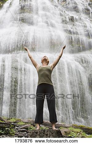 Woman standing with arms raised at the foot of a waterfall View Large Photo Image Stock Photograph - Woman standing with arms raised at the foot of a waterfall. Fotosearch