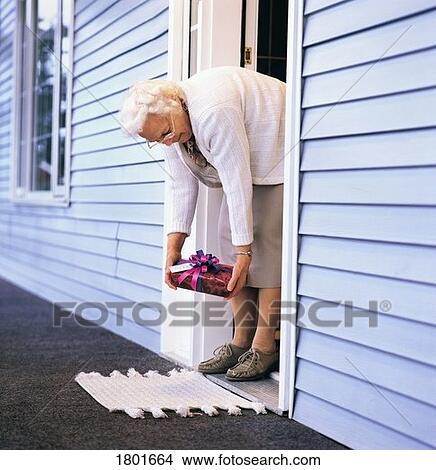 Picture - Elderly woman picking up a gift from her front doorway. Fotosearch