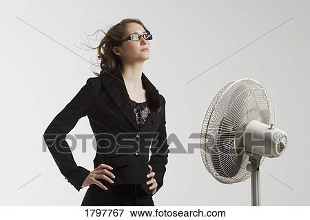 Stock Photo - Young woman standing in front of an electric fan. Fotosearch
