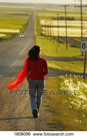 Stock Photograph - Woman walking along highway surrounded by fields. Fotosearch