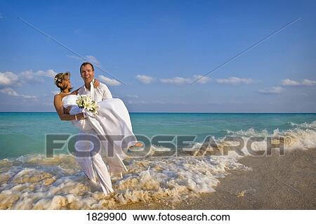 Stock Image - Groom carrying bride on beach. Fotosearch