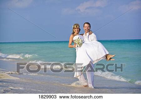 Stock Image - Groom carrying bride on beach. Fotosearch