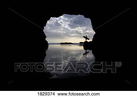 Picture - Silhouette of surfer on beach through cave, Muriwai Beach, North Island, New Zealand. Fotosearch