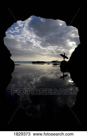 Stock Image - Silhouette of surfer on beach through cave, Muriwai Beach, North Island, New Zealand. Fotosearch