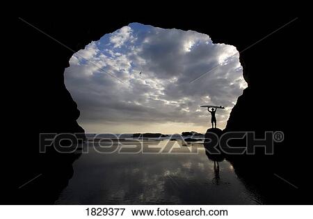 Stock Photo - Silhouette of surfer on beach through cave, Muriwai Beach, North Island, New Zealand. Fotosearch