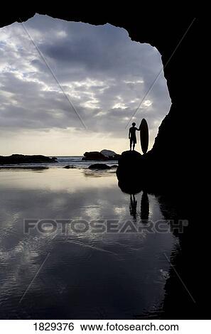 Stock Photograph - Silhouette of surfer on beach through cave, Muriwai Beach, North Island, New Zealand. Fotosearch