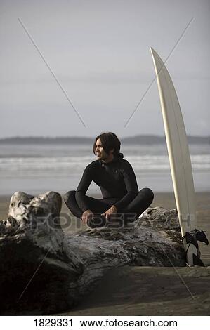 Stock Image - Surfer sitting on log on beach, Cox Bay near Tofino, British Columbia, Canada. Fotosearch