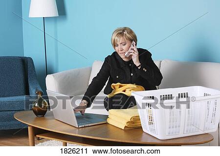 Stock Image - Woman on the phone, working on her laptop and folding laundry. Fotosearch