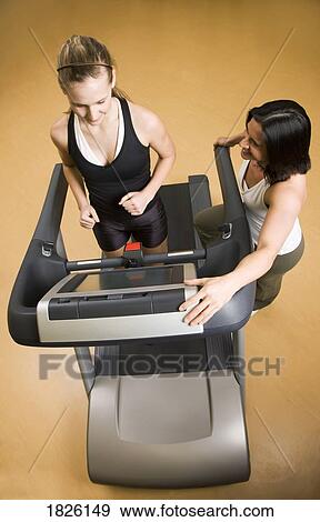 Stock Photo - Woman running on the treadmill with other woman watching. Fotosearch