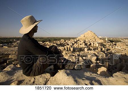 Stock Image - Man overlooking town, Siwa Town, Egypt; man sitting on top of ancient fortress overlooking town. Fotosearch