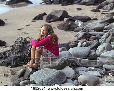 Stock Image - Maui, Hawaii, USA; Young girl sitting on some rocks near the water. Fotosearch