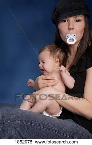 Stock Photography - Woman and baby; Young woman holding infant in her lap with pacifier in her mouth. Fotosearch