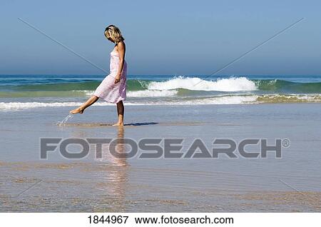 Stock Photo - Young woman walking along the beach kicking up water. Fotosearch
