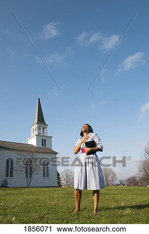 Stock Image - Woman outside with Bible, standing near a church, praying. Fotosearch