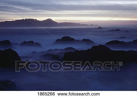 Stock Photograph - Fog around coastal rocks at twilight. Fotosearch