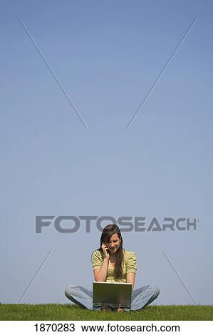 Stock Image - a girl working on a laptop and talking on a cellphone while sitting on the grass. Fotosearch