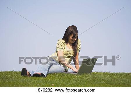 Stock Image - a girl working on her laptop while sitting on the grass. Fotosearch