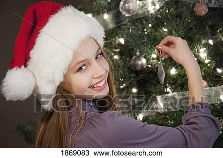 Stock Image - girl decorating the christmas tree wearing a santa hat. Fotosearch