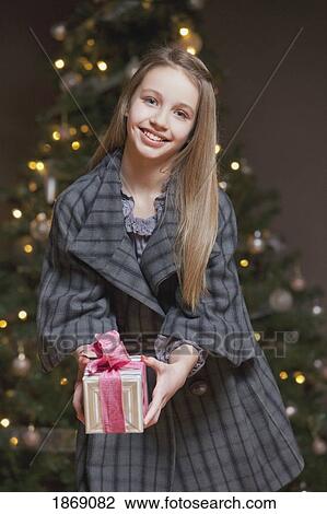 girl holding a gift standing in front of the christmas tree View Large Photo Image Stock Image - girl holding a gift standing in front of the christmas tree. Fotosearch