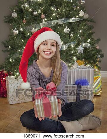 Stock Image - girl sitting in front of the christmas tree holding a gift and wearing a santa hat. Fotosearch