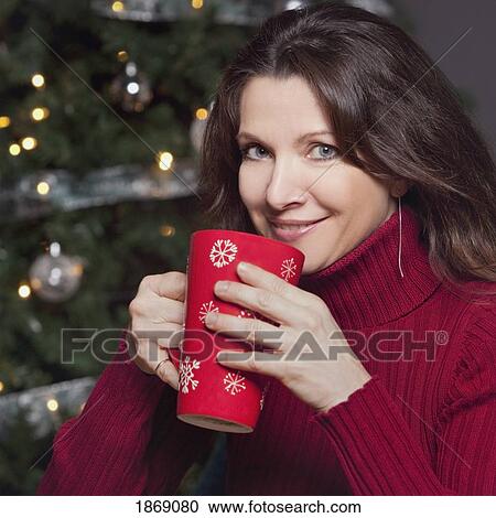 Stock Image - woman sipping a hot beverage by the christmas tree. Fotosearch