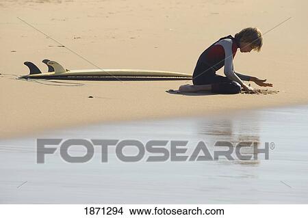 Picture - a boy playing in the sand and sitting beside his surfboard. Fotosearch
