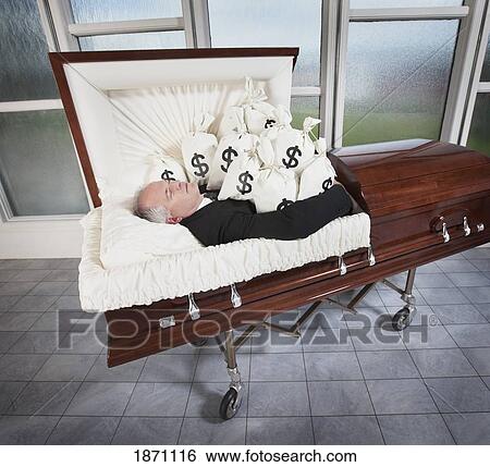 Stock Photograph - a man laying in a coffin surrounded by bags of money. Fotosearch