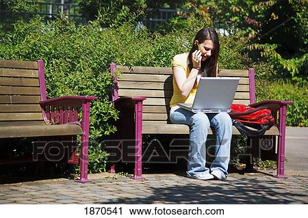 a young woman working on her laptop computer and talking on her cellphone while sitting outdoors on a park bench View Large Photo Image Stock Image - a young woman working on her laptop computer and talking on her cellphone while sitting outdoors on a park bench. Fotosearch