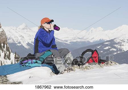 Stock Image - banff national park, alberta, canada; a female hiker sits to rest and take a drink and is surrounded by snow-covered mountains. Fotosearch