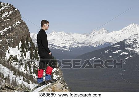 lake louise, banff national park, alberta, canada; a young man standing on a rock ledge overlooking snow-covered mountains and a valley View Large Photo Image Stock Photo - lake louise, banff national park, alberta, canada; a young man standing on a rock ledge overlooking snow-covered mountains and a valley. Fotosearch