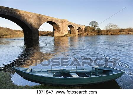Stock Photo - northumberland, england; a boat moored on the edge of a river with a bridge. Fotosearch