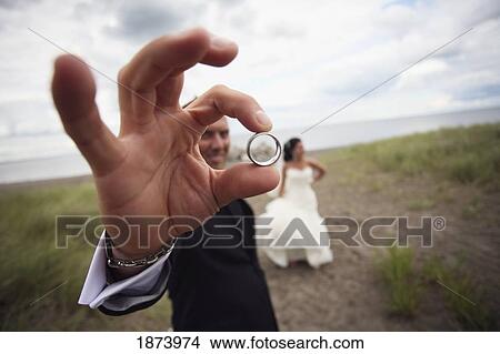 Picture - a bride and groom standing on a dirt road and the groom is holding his wedding band. Fotosearch