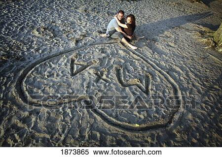 Stock Photography - a couple sitting in the sand together under a heart with their initials. Fotosearch
